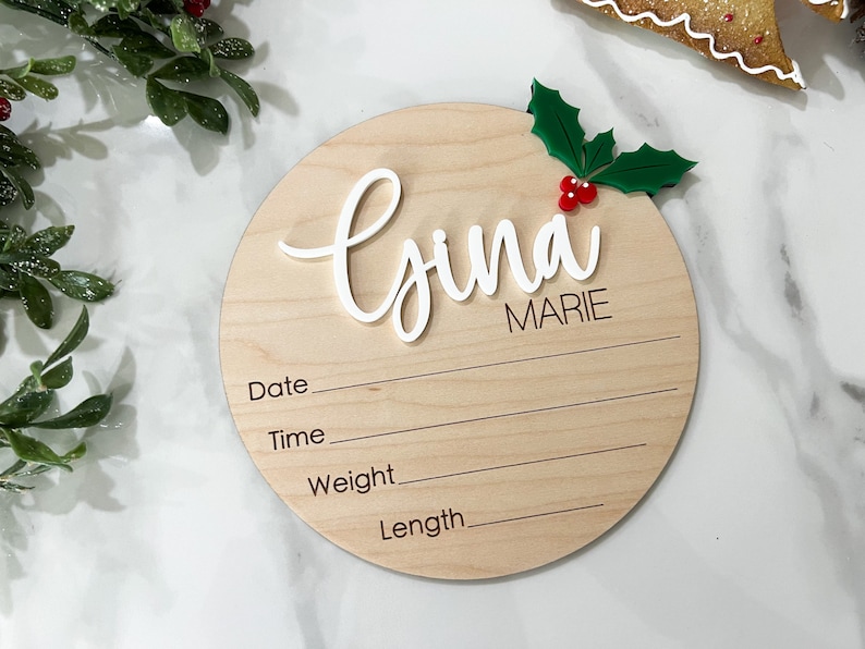 May include: A round wooden plaque with a white cursive name "Gina Marie" and a holly sprig with red berries. The plaque has lines for recording the date, time, weight, and length.