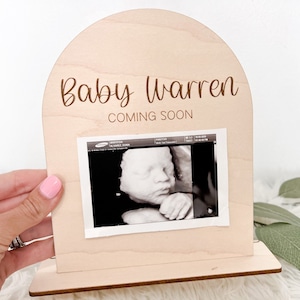 May include: A wooden arch-shaped sign with the text "Baby Warren Coming Soon" and a black and white ultrasound image.