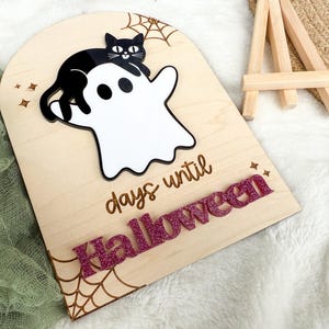 K&ouml;nnte beinhalten: H&ouml;lzernes Halloween-Countdown-Schild mit einer schwarzen Katze auf einem wei&szlig;en Geist. Das Schild zeigt "Tage bis Halloween" mit glitzernden rosa Buchstaben. Ein Spinnennetz-Design ist auf das Holz ge&auml;tzt.