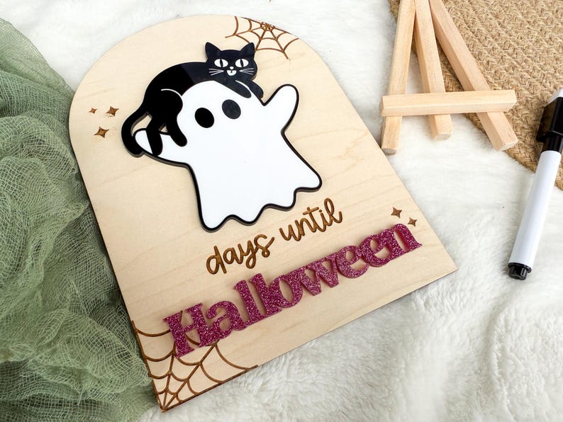 K&ouml;nnte beinhalten: H&ouml;lzernes Halloween-Countdown-Schild mit einer schwarzen Katze auf einem wei&szlig;en Geist. Das Schild zeigt "Tage bis Halloween" mit glitzernden rosa Buchstaben. Ein Spinnennetz-Design ist auf das Holz ge&auml;tzt.