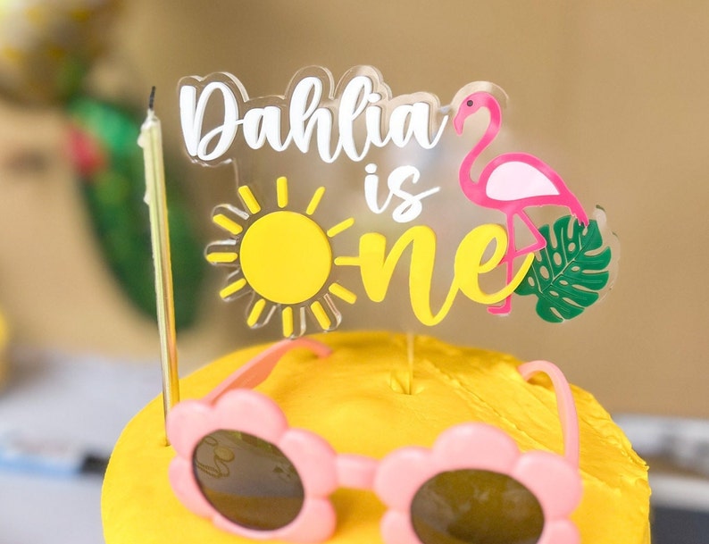 May include: A yellow cake with a pink flamingo, a yellow sun, and the words "Dahlia is one" on a clear acrylic topper. The cake is decorated with pink sunglasses.