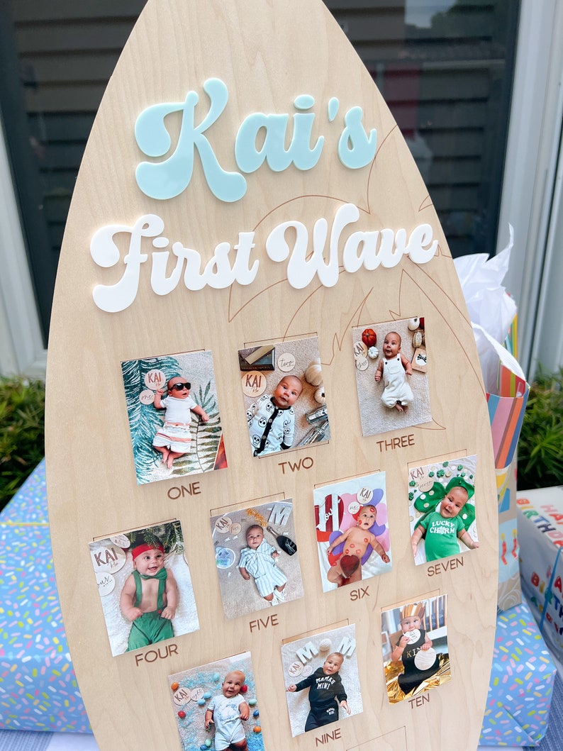 May include: A wooden surfboard-shaped sign with the text "Kai's First Wave" in white and blue letters. The sign has ten square photo frames with pictures of a baby boy at different ages, each frame labeled with a number from one to ten.