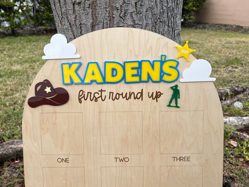 Peut inclure: Un panneau en bois clair avec un sommet arqu&eacute; portant le texte KADEN'S first round up en bleu, jaune et marron. Les &eacute;l&eacute;ments d&eacute;coratifs incluent des nuages blancs, un chapeau de cowboy marron, une &eacute;toile de sh&eacute;rif jaune et un soldat vert.