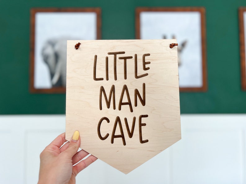 May include: A wooden sign with the words "LITTLE MAN CAVE" carved into the surface. The sign is light-colored with a pointed bottom and two small holes at the top. The sign is being held in front of a green wall.