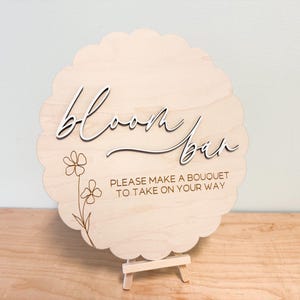 Pu&ograve; includere: Un cartello di legno con un bordo a festoni e un design floreale. Il cartello dice "Bloom Bar" e "Per favore, fai un bouquet da portare via."