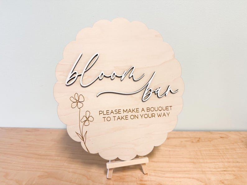 Pu&ograve; includere: Un cartello di legno con un bordo a festoni e un design floreale. Il cartello dice "Bloom Bar" e "Per favore, fai un bouquet da portare via."