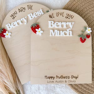 May include: Two wooden arch-shaped signs with the phrases "You Are The Berry Best" and "We Love You Berry Much" in white lettering. Each sign has strawberry and flower embellishments. The bottom sign reads "Happy Mother's Day! Love, Austin & Olivia."
