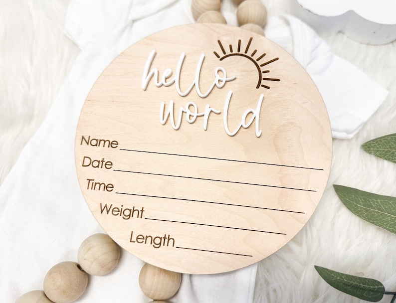 May include: A round wooden sign with the words "hello world" in white script. The sign has a brown sun design and blank lines for name, date, time, weight, and length.