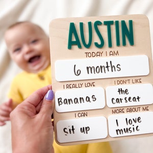 May include: A rectangular wooden milestone board with the name AUSTIN in dark green raised letters. The board features white panels with black text reading: TODAY I AM 6 months, I REALLY LOVE Bananas, I DON'T LIKE the carseat, I CAN sit up, and MORE ABOUT ME I love music.