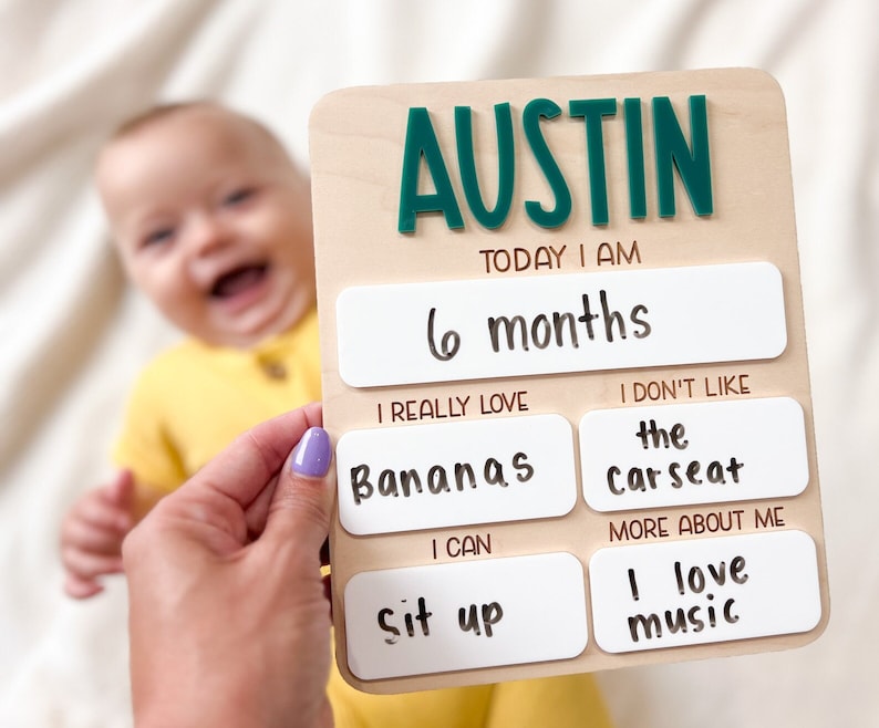 May include: A rectangular wooden milestone board with the name AUSTIN in dark green raised letters. The board features white panels with black text reading: TODAY I AM 6 months, I REALLY LOVE Bananas, I DON'T LIKE the carseat, I CAN sit up, and MORE ABOUT ME I love music.