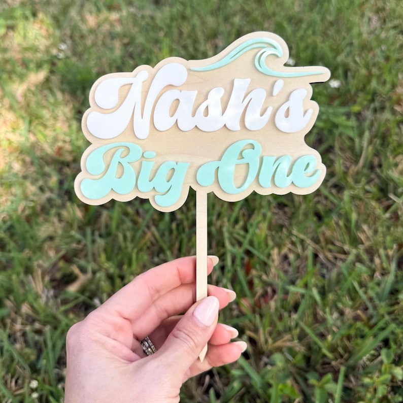 May include: A wooden cake topper with the text "Nash's Big One" in white and teal lettering. The topper has a wave design at the top and is held by a wooden stick. The background is green grass.