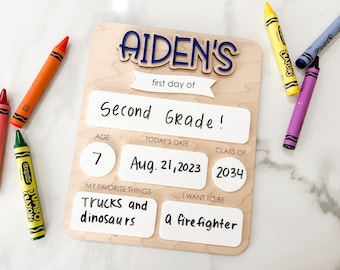 May include: A wooden sign with the name "Aiden's" in blue letters, a white banner that says "first day of", and a white rectangle that says "Second Grade!" The sign also has sections for age, today's date, class of, favorite things, and what the child wants to be when they grow up. The sign has the numbers 7, 2034, and the date August 21, 2023 filled in. The favorite things section says "Trucks and dinosaurs" and the "I want to be" section says "a firefighter".