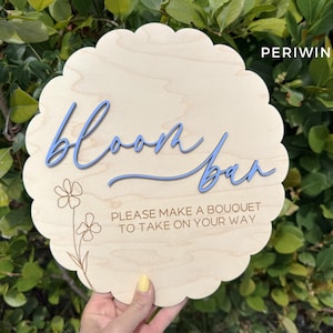 Pu&ograve; includere: Un cartello di legno con un bordo a festoni e il testo "Bloom Bar" in lettere blu. Il cartello include anche il testo "Please make a bouquet to take on your way" in lettere marroni.