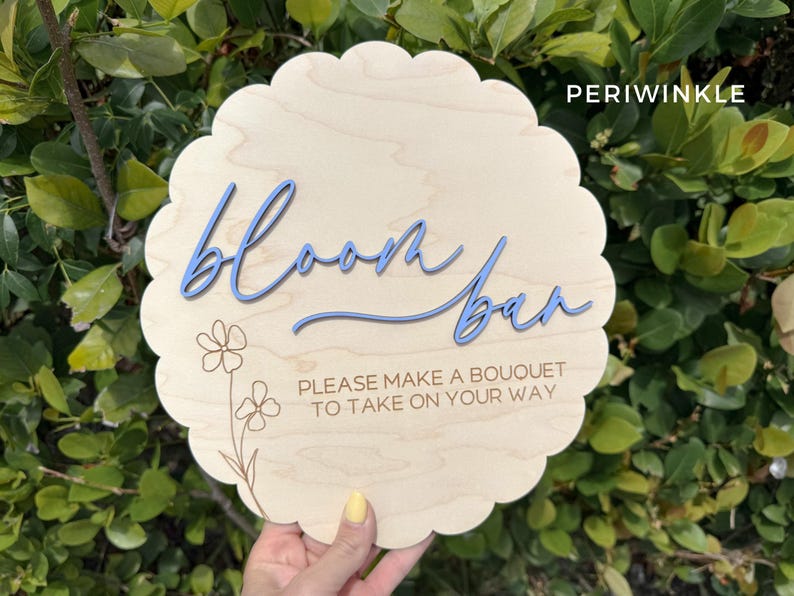 Pu&ograve; includere: Un cartello di legno con un bordo a festoni e il testo "Bloom Bar" in lettere blu. Il cartello include anche il testo "Please make a bouquet to take on your way" in lettere marroni.