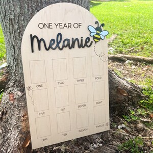 Op de afbeelding: Een boogvormig houten mijlpaalbord met de tekst ONE YEAR OF Melanie in zwarte letters, een bijengrafiek en twaalf rechthoekige fotovakken gelabeld met de cijfers &eacute;&eacute;n tot en met twaalf in zwarte tekst op een licht houten oppervlak.