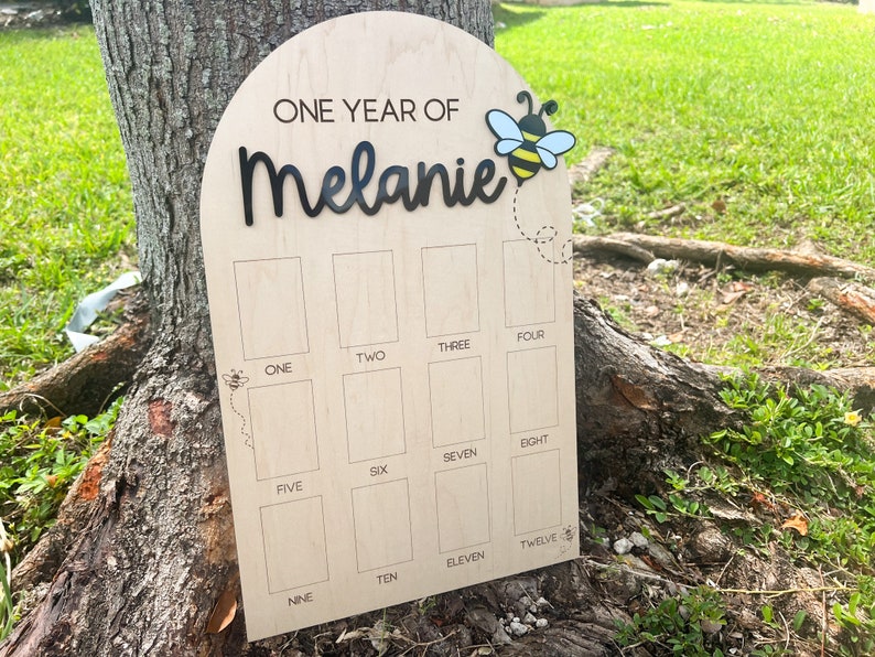Op de afbeelding: Een boogvormig houten mijlpaalbord met de tekst ONE YEAR OF Melanie in zwarte letters, een bijengrafiek en twaalf rechthoekige fotovakken gelabeld met de cijfers &eacute;&eacute;n tot en met twaalf in zwarte tekst op een licht houten oppervlak.