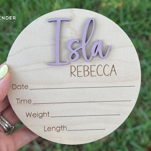 May include: A round wooden baby announcement sign with the name "Isla" in lavender script and the name "Rebecca" in a simple font. The sign includes lines for date, time, weight, and length. The word "Lavender" is in the top left corner.
