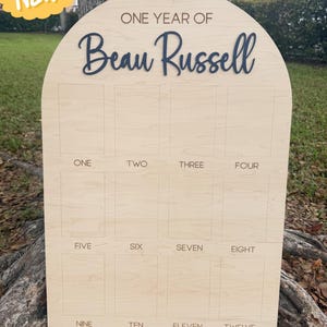 Wooden arch photo board with the text "ONE YEAR OF Beau Russell" in a dark blue font. The board has 12 photo slots labeled "ONE" through "TWELVE". The board is 21.2 x 30.3 inches.  This birthday boar is for 4x6' photos.