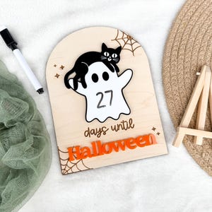 K&ouml;nnte beinhalten: Ein h&ouml;lzernes Halloween-Countdown-Schild mit einer schwarzen Katze auf einem wei&szlig;en Geist, der die Zahl 27 anzeigt. Die Worte "days until Halloween" stehen darunter, mit orangefarbenen Buchstaben, die "Halloween" unten buchstabieren. Ein wei&szlig;er Marker und eine kleine Holzstaffelei sind ebenfalls vorhanden.