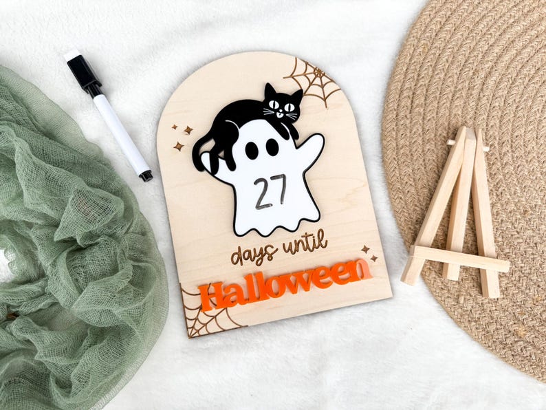 K&ouml;nnte beinhalten: Ein h&ouml;lzernes Halloween-Countdown-Schild mit einer schwarzen Katze auf einem wei&szlig;en Geist, der die Zahl 27 anzeigt. Die Worte "days until Halloween" stehen darunter, mit orangefarbenen Buchstaben, die "Halloween" unten buchstabieren. Ein wei&szlig;er Marker und eine kleine Holzstaffelei sind ebenfalls vorhanden.