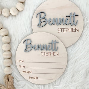 K&ouml;nnte beinhalten: Zwei runde Holzschilder mit dem Namen "Bennett" in grauer Schrift und dem Namen "Stephen" darunter. Die Schilder haben Linien f&uuml;r Datum, Uhrzeit, Gewicht und L&auml;nge. Auf einem wei&szlig;en, flauschigen Hintergrund.