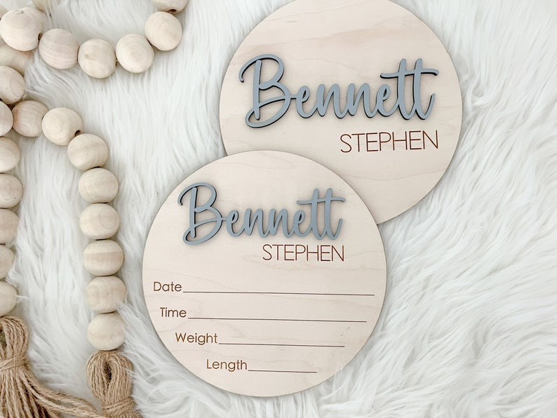 K&ouml;nnte beinhalten: Zwei runde Holzschilder mit dem Namen "Bennett" in grauer Schrift und dem Namen "Stephen" darunter. Die Schilder haben Linien f&uuml;r Datum, Uhrzeit, Gewicht und L&auml;nge. Auf einem wei&szlig;en, flauschigen Hintergrund.