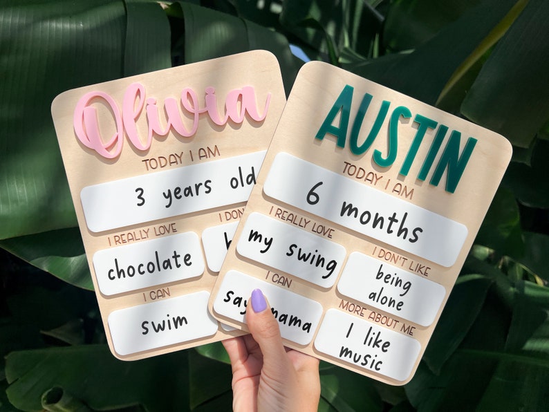 May include: Two light-colored wooden milestone boards with rounded corners. One board features the name Olivia in pink, the other Austin in green. Both include white rectangular sections with printed text detailing age, likes, dislikes, and abilities.