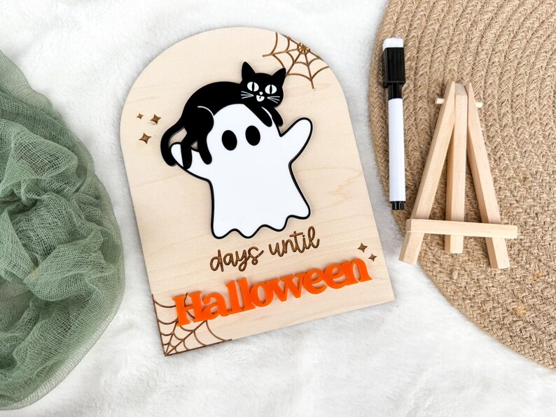 K&ouml;nnte beinhalten: H&ouml;lzernes Halloween-Countdown-Schild mit einem wei&szlig;en Geist und einer schwarzen Katze. Das Schild enth&auml;lt den Text "days until Halloween" und orangefarbene Akzente. Ein schwarzer Marker und eine kleine Holzstaffelei sind ebenfalls vorhanden.