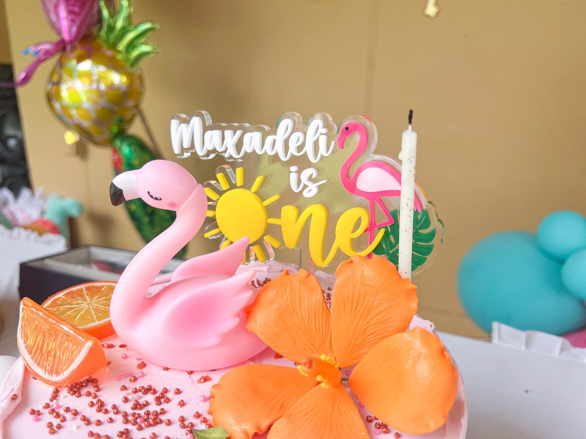 May include: A pink and white cake with a flamingo cake topper, a sun, and the words "Maxadeli is one". The cake is decorated with orange flowers, orange slices, and pink sprinkles.