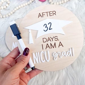 May include: A wooden round sign with the text "After 32 days, I am a NICU grad" in white letters. A white graduation cap is on the sign. A black dry-erase marker is next to the sign.