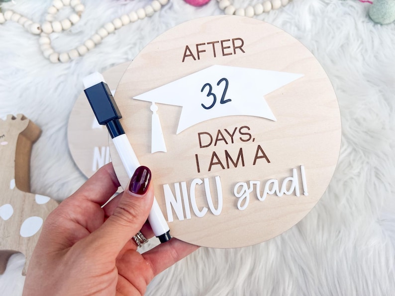 May include: A wooden round sign with the text "After 32 days, I am a NICU grad" in white letters. A white graduation cap is on the sign. A black dry-erase marker is next to the sign.