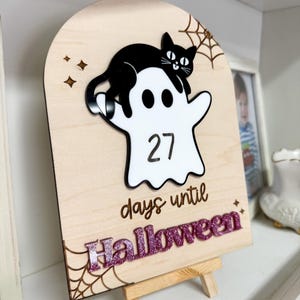 K&ouml;nnte beinhalten: H&ouml;lzernes Halloween-Countdown-Schild mit einem wei&szlig;en Geist und einer schwarzen Katze. Das Schild zeigt "27 Tage bis Halloween" in einer glitzernden lila Schrift. Ein Spinnennetz-Design befindet sich in der Ecke.