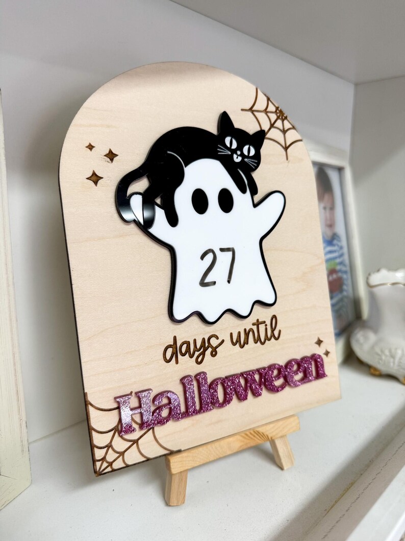 K&ouml;nnte beinhalten: H&ouml;lzernes Halloween-Countdown-Schild mit einem wei&szlig;en Geist und einer schwarzen Katze. Das Schild zeigt "27 Tage bis Halloween" in einer glitzernden lila Schrift. Ein Spinnennetz-Design befindet sich in der Ecke.