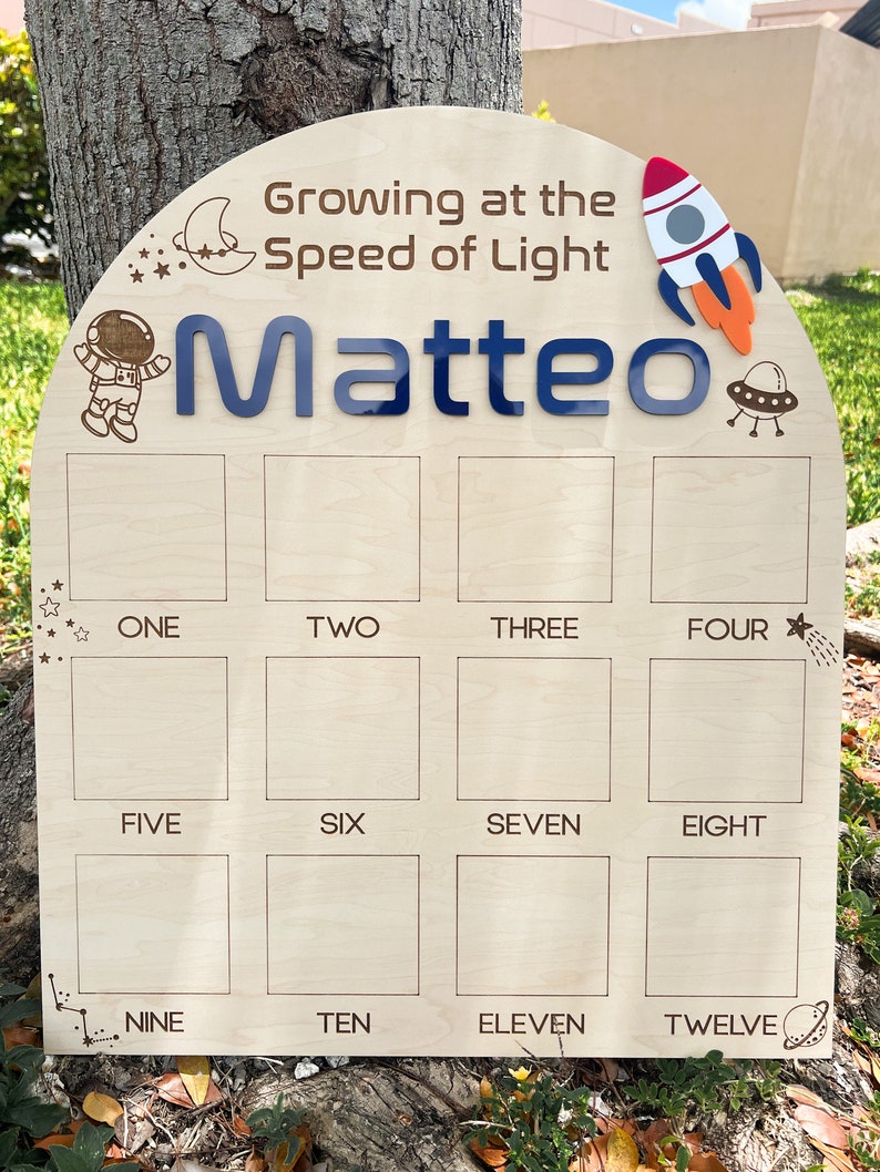 Pu&ograve; includere: Un grafico di crescita in legno a tema spaziale. Presenta un razzo, un astronauta, una luna, stelle e pianeti. Il grafico ha 12 quadrati vuoti per registrare le misure di altezza. Il testo "Growing at the Speed of Light" e "Matteo" sono stampati sul grafico.