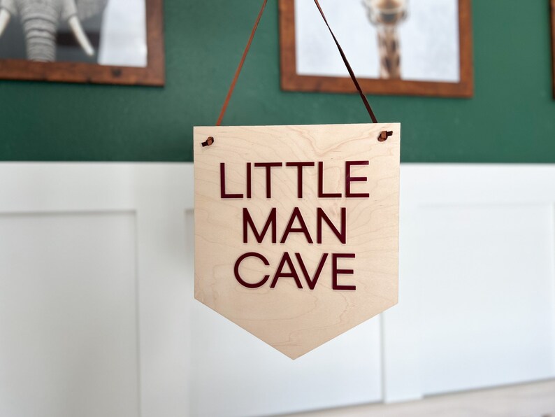 May include: A wooden sign with the words "LITTLE MAN CAVE" in dark red lettering. The sign is a light wood color and is hanging from a brown ribbon. The sign is in a room with framed pictures on the wall.