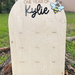 Op de afbeelding: Een boogvormig houten mijlpaalbord met de tekst ONE YEAR OF Kylie in zwart en bruin. Het bord bevat twaalf vierkante fotokaders gelabeld van ONE tot TWELVE, versierd met gegraveerde bijenillustraties en een 3D-bijenaccent.