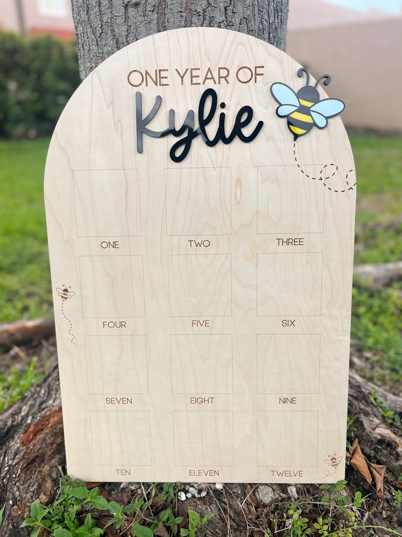 Op de afbeelding: Een boogvormig houten mijlpaalbord met de tekst ONE YEAR OF Kylie in zwart en bruin. Het bord bevat twaalf vierkante fotokaders gelabeld van ONE tot TWELVE, versierd met gegraveerde bijenillustraties en een 3D-bijenaccent.