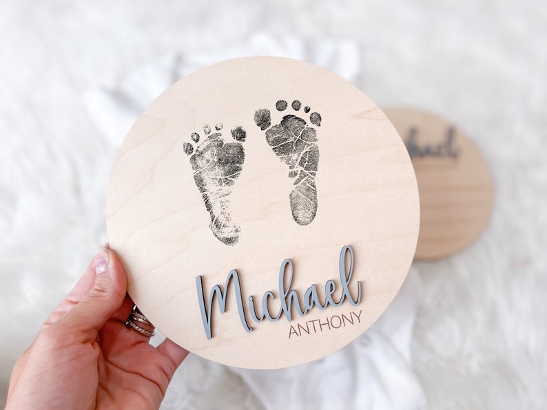 May include: A round wooden plaque with two baby footprints in black ink. The name "Michael Anthony" is displayed in gray, cursive lettering. A second plaque is visible in the background.