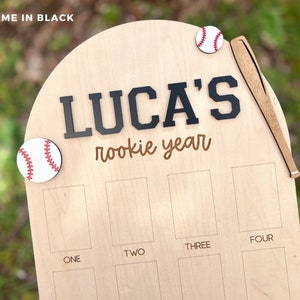 May include: A light-colored wooden arched plaque featuring the text LUCA'S rookie year in black and brown lettering. The plaque is decorated with two circular baseball icons and a wooden baseball bat. Below, there are eight rectangular slots labeled ONE, TWO, THREE, and FOUR.
