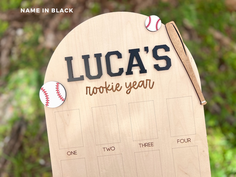 May include: A light-colored wooden arched plaque featuring the text LUCA'S rookie year in black and brown lettering. The plaque is decorated with two circular baseball icons and a wooden baseball bat. Below, there are eight rectangular slots labeled ONE, TWO, THREE, and FOUR.