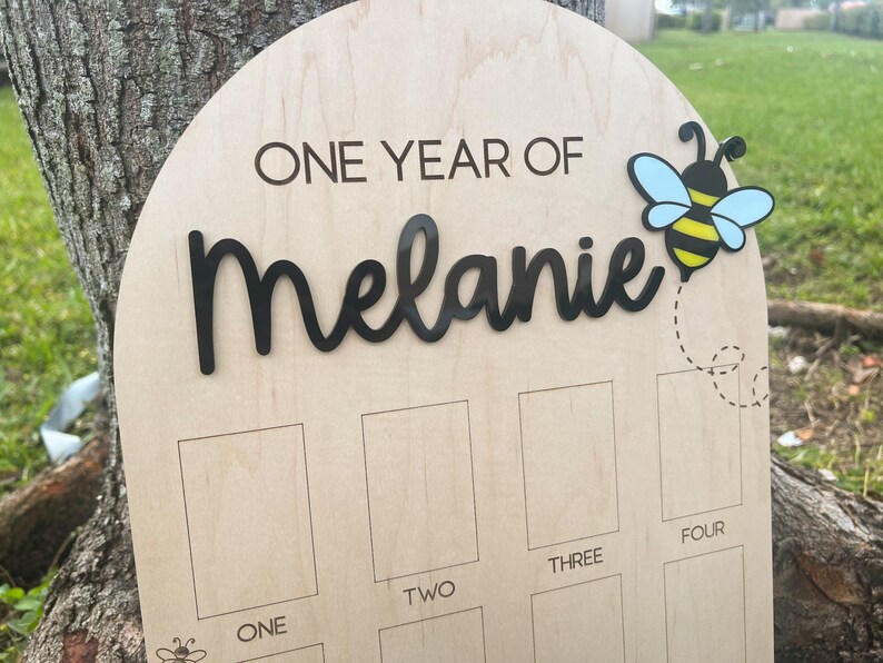 Op de afbeelding: Een boogvormig houten mijlpaalbord met de tekst ONE YEAR OF Melanie in zwarte letters. Een bijengrafiek met blauwe vleugels en gele en zwarte strepen is rechtsboven bevestigd. Daaronder bevinden zich vier rechthoekige fotokaders met de labels ONE, TWO, THREE en FOUR.