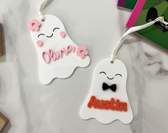 Op de afbeelding: Twee witte, spookvormige ornamenten met roze en oranje letters. Het eerste spook heeft roze bloemen en de naam "Olivia" in roze geschreven. Het tweede spook heeft een zwarte strik en de naam "Austin" in oranje geschreven.