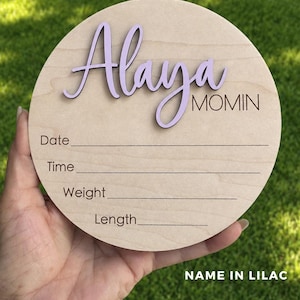 May include: A circular wooden plaque featuring the name Alaya in raised lilac script lettering and the word MOMIN in black text. Below, the plaque includes engraved lines with the labels Date, Time, Weight, and Length for recording details.