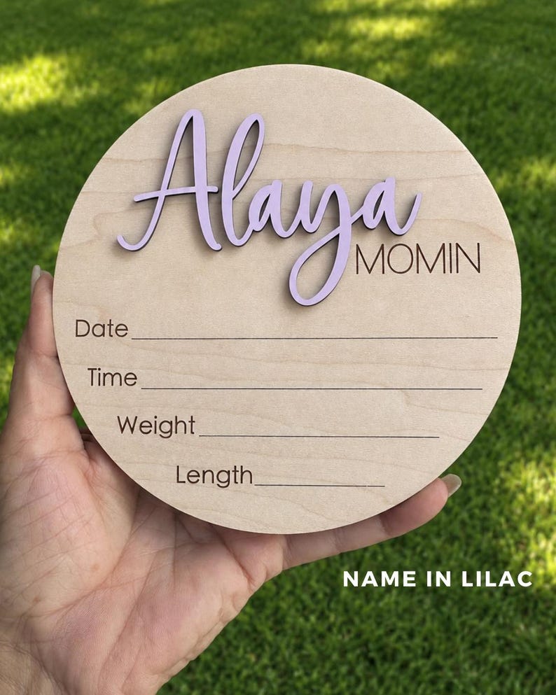 May include: A circular wooden plaque featuring the name Alaya in raised lilac script lettering and the word MOMIN in black text. Below, the plaque includes engraved lines with the labels Date, Time, Weight, and Length for recording details.