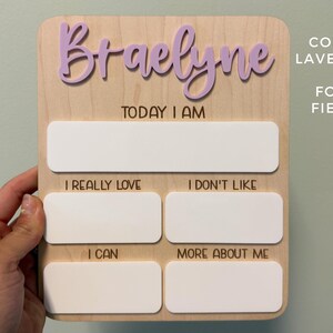 May include: A rectangular wooden milestone board featuring the name Braelyne in raised lavender script lettering. The board includes the text Today I Am, I Really Love, I Don't Like, I Can, and More About Me, with blank white spaces for writing.