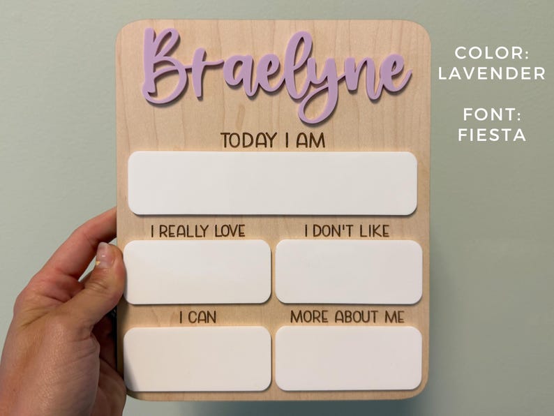 May include: A rectangular wooden milestone board featuring the name Braelyne in raised lavender script lettering. The board includes the text Today I Am, I Really Love, I Don't Like, I Can, and More About Me, with blank white spaces for writing.