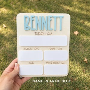 May include: A rectangular wooden milestone board featuring the name BENNETT in raised light blue letters. The board includes white dry-erase sections labeled TODAY I AM, I REALLY LOVE, I DON'T LIKE, I CAN, and MORE ABOUT ME for tracking developmental milestones.