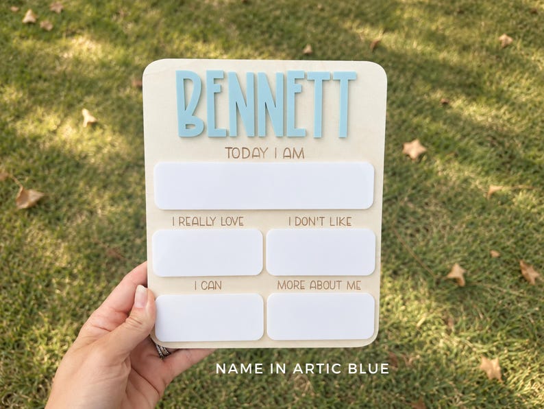 May include: A rectangular wooden milestone board featuring the name BENNETT in raised light blue letters. The board includes white dry-erase sections labeled TODAY I AM, I REALLY LOVE, I DON'T LIKE, I CAN, and MORE ABOUT ME for tracking developmental milestones.