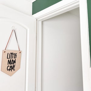 May include: A wooden sign with the text "LITTLE MAN CAVE" in black lettering hangs on a white door. The sign is a light brown color and is suspended by a brown and black cord. The door is slightly ajar, revealing a white interior.