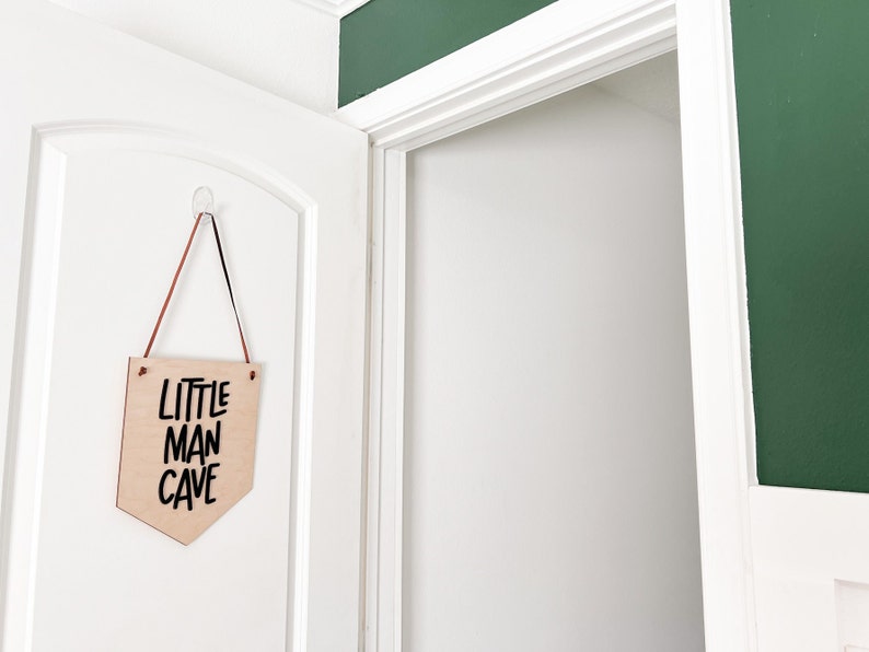 May include: A wooden sign with the text "LITTLE MAN CAVE" in black lettering hangs on a white door. The sign is a light brown color and is suspended by a brown and black cord. The door is slightly ajar, revealing a white interior.